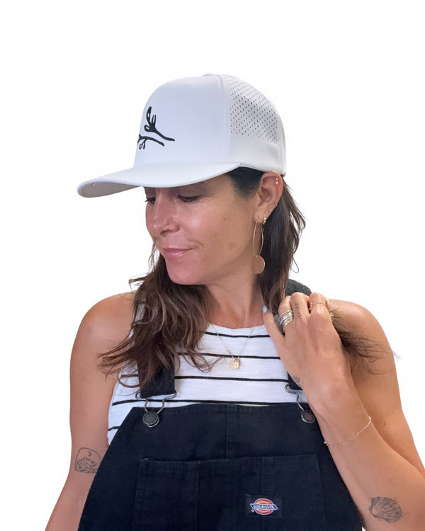 Woman wearing a white Agapantha cap with a logo, striped tank top, and black overalls on a white background